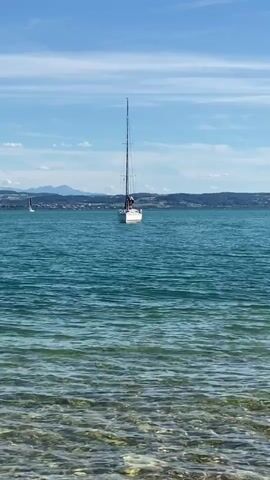 The video displays a wide shot of a clear blue sky with sparse white clouds above a tranquil body of water. In the foreground, the shallow water reveals a rocky seabed, with sunlight creating shimmering patterns on the surface. Further out, a white sailboat with a tall mast is anchored, with one person visible on deck. In the distance, hazy landmasses and mountains can be seen under the bright sky. Another, smaller sailboat is visible further to the left. The overall scene conveys a peaceful, sunny day by the water.