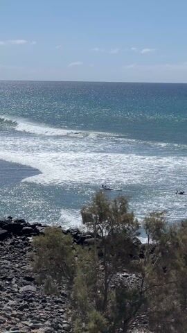 The video shows a few surfers paddling into waves in the ocean. The water is a blue-green color and the sky is mostly blue with some clouds. There are some trees and rocks in the foreground. The surfers are spread out, paddling into the surf. The waves are relatively small. The video is shot from an elevated position, providing a clear view of the ocean and the surfers.