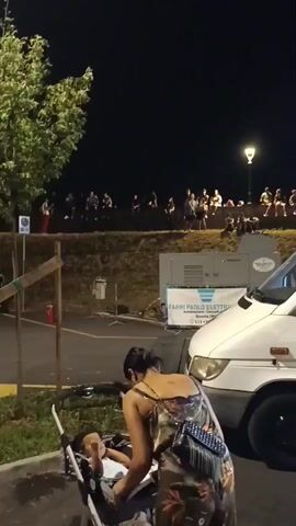 The video is taken at night, outdoors, likely at a food truck festival or outdoor market. In the foreground, a woman wearing a floral dress is attending to a baby in a stroller. To the right, a white food truck is visible with its serving window open. Inside the food truck, at least two people appear to be working, preparing food. Several customers are standing in line or at the serving window of the food truck. Further in the background, there are more food stalls or tents, with people milling about. The overall atmosphere suggests a lively evening event. The lighting is a mix of artificial lights from the food trucks and stalls, and ambient night light. There are trees and some low walls visible in the background, and a street lamp can be seen in the distance. A white van is parked next to the food truck. The ground appears to be paved, possibly a parking lot.
