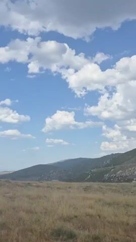 A vertical video opens on a landscape with a clear blue sky dotted with white, fluffy clouds. In the distance, rolling hills covered in green vegetation and rocky outcrops are visible. The foreground consists of dry, golden grass and patches of dirt. As the video progresses, a black and yellow striped pole enters the frame from the right, moving across the view. The camera continues to move, showing more of the landscape and the pole momentarily obstructing the view. The pole then exits the frame to the left, and the video concludes with the same landscape view as the beginning.