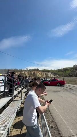 The video shows a group of people watching a red car perform a drift on a racetrack. The people are standing on a metal platform behind a metal railing. The sky is blue with some clouds. Some people are holding their phones, possibly recording the car's drift. There is a dirt hill in the background with some green trees. The car is in the middle of the track, drifting sideways. The scene appears to be a motorsports event.