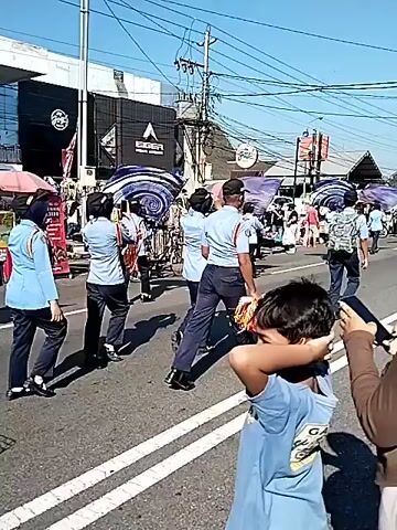 A medium shot captures a sunny outdoor street scene with multiple people and buildings. In the foreground, the back of a person with a light brown head covering and a person holding a child face the viewer. In the midground, several individuals in light blue shirts and dark pants walk down a street. One person cycles past them on a white bicycle. The background reveals buildings, power lines, and market stalls with colorful umbrellas. The road has white double lines. The light is bright, suggesting daytime.