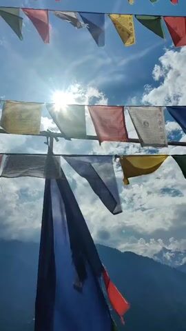 The video shows prayer flags strung across a bright, sunny sky with clouds. The flags are of various colors, including red, blue, yellow, green, and white, and some appear to have writing on them. The sun is shining brightly, creating a lens flare effect. In the background, there are mountains and a valley with buildings. The viewpoint is from below, looking up at the flags and the sky.