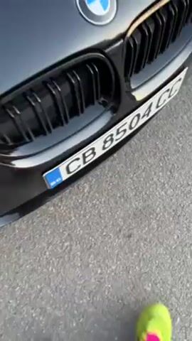 The video shows a black BMW car. The camera starts close-up on the front grill and license plate, then pulls back to show the front of the car. The license plate is partially visible and appears to read "CB 8504C." A person wearing a bright green shoe stands in front of the car, and their hand briefly appears to push the camera away. The environment appears to be an outdoor parking area with asphalt.