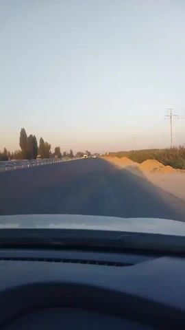 The video shows a view from inside a vehicle traveling on a highway. The time stamp indicates it is from 00:00 to 00:04. The road appears to be under construction or has recently undergone repairs, with dirt piles visible on the right side of the road. Several vehicles, including cars and trucks, are on the highway in front of and beside the filming vehicle. The sky is clear and the lighting suggests it is either early morning or late afternoon. Trees and power lines are visible along the side of the road.