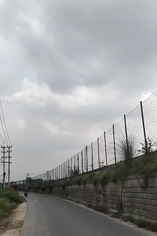 The video shows a two-lane road under a cloudy sky. A motorcycle drives on the road. A long retaining wall runs alongside the road, topped with a tall chain-link fence. Utility poles are visible on the left side of the road. The overall scene appears somewhat industrial or suburban.