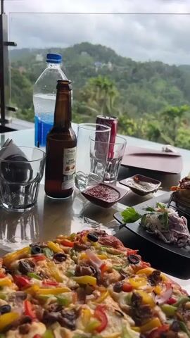 A video shows a table laden with food and drinks. The table is made of glass, reflecting the items on it. In the foreground, a large, colorful pizza with various toppings is visible. To the right of the pizza, there are several small black plates holding different dishes, including what appears to be a salad or coleslaw and some other appetizers. Several small bowls contain sauces or dips. A brown bottle, possibly of beer, and a plastic bottle of water are on the table. Two empty drinking glasses are also present. A red can, possibly a soda, is next to one of the glasses. In the background, a stunning panoramic view of lush green mountains covered with trees and some scattered buildings under a cloudy sky is visible through a glass barrier. The camera slightly pans to the right, showing more of the mountainous landscape.