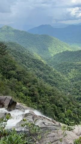 The video shows a view from the top of a waterfall overlooking a lush, green valley. The waterfall cascades over rocks on the left side of the frame, with white water rushing over them. The valley is filled with dense trees that cover rolling hills and mountains that extend into the distance. The sky is cloudy with patches of sunlight breaking through, illuminating parts of the landscape. The overall scene is one of natural beauty and serenity. There are no people or man-made objects visible in the video.