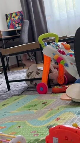 The video shows a baby lying on their back under a table. The baby is wearing light-colored pajamas with a teddy bear pattern. A walker toy with bright orange legs, a green handle, and white and colorful interactive buttons is positioned next to the baby, partially under the table. The baby's legs are visible, extended towards the walker. A black and white patterned toy hangs from the table above the baby. The floor is covered with a large, colorful play mat with a road or city design. A grey rug is visible beneath the play mat. In the background, a chair and a framed colorful artwork are present. The scene appears to be indoors, possibly a living room or playroom, with natural light coming from a window covered by a grey curtain. The video captures a moment of the baby interacting with or near the walker toy.