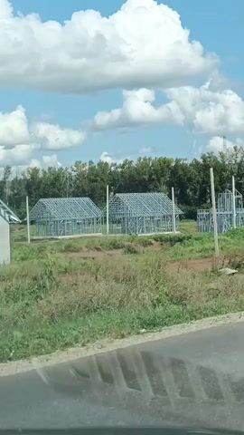 The video shows a construction site with several unfinished houses. The houses are framed with metal structures, likely light gauge steel. Two of the houses are further along, with walls and roofs partially completed. The site is surrounded by trees and a grassy field. The sky is blue with fluffy white clouds. The video appears to be shot from a moving vehicle, possibly a car, as indicated by the reflection on the bottom of the screen.