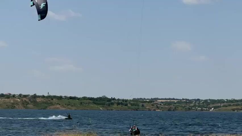 The video shows a bright, sunny day with a clear blue sky and scattered clouds. A body of water, likely a lake or bay, fills the lower half of the frame. In the distance, a green, hilly shoreline is visible, dotted with houses and trees, suggesting a coastal or lakeside town. The water itself is dark blue with small ripples, indicating a light breeze.

The main action involves two individuals engaged in kitesurfing. In the foreground, a person on a board is being pulled by a large, black and blue kite with a white star. This individual is positioned closer to the viewer and is actively maneuvering through the water, leaving a white wake behind them.

Further in the middle ground, another kitesurfer is visible, also on a board and connected to a kite. This second kitesurfer appears to be further from the shore and also engaged in the activity. The kites are high in the sky, tethered to the surfers by thin lines. The overall scene depicts a recreational water sport taking place in a scenic, natural environment.