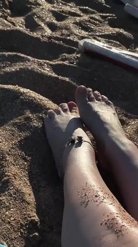 The video shows a close-up of a person's feet partially covered in wet sand on a beach. An anklet with a charm is visible on one foot. The camera pans slightly to reveal the ocean and the horizon in the background, with several people in silhouette sitting or standing on the beach in the distance. The lighting suggests it is daytime, possibly late afternoon. The overall environment is a beach with sand and water.
