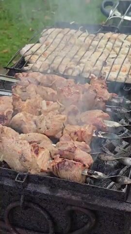A close-up shot shows skewers of meat cooking over hot coals on a grill. Above the skewers, another grill holds numerous pieces of pale meat, possibly chicken or fish. Smoke rises from the cooking meat. The grill appears to be outdoors, with a hint of green grass in the background. The scene captures the process of barbecuing.