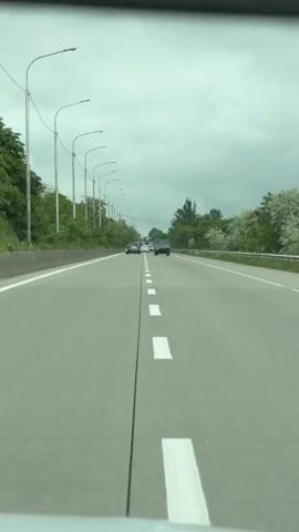 The video shows a car driving on a highway. There are two cars in front. There are street lights on the side of the road. The sky is cloudy. There are trees on both sides of the road.