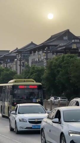 The video shows a street in China. A yellow and black bus is stopped in traffic, behind a white Chevrolet Malibu. A scooter with two men wearing casual clothes passes by on the left. The street is lined with buildings in a traditional Chinese architectural style, and some green trees. The sky is hazy, and the sun is a bright white disk in the sky.