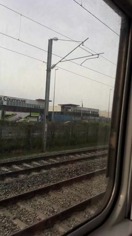 The view from a train window shows two sets of train tracks covered in ballast. Beyond the tracks is a low green hedge. Behind the hedge is a large grey building with a red section on the roof. Graffiti covers a wall on the building.  Several overhead power lines stretch across the sky. A light post can be seen in the distance. The sky is overcast.