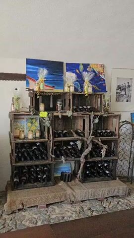 The video shows a display of wine bottles and jars of food items. The display is arranged on wooden crates. Two paintings hang on the wall behind the display. The room appears to be a shop or tasting room. There are no people visible in the video.