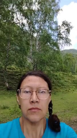 A selfie video opens on a woman wearing glasses and a teal shirt. Her hair is dark and styled into a braid. She is standing in an outdoor setting with trees and grass visible in the background. The camera moves slightly, revealing more of the wooded area and a gently sloping hillside covered in grass. In the distance, two dark figures, possibly cattle, can be seen grazing. The video ends with the camera still focused on the woman.