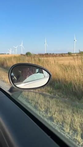 A person is visible in the reflection of a car's side mirror. They are looking into a phone and smiling. The car is moving, as indicated by the blurred background of a field of dry grass and wind turbines under a clear blue sky. The perspective is from inside the car, looking out the window and at the side mirror.