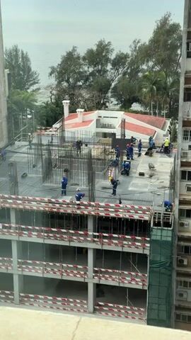 The video shows a construction site. Several construction workers, wearing blue shirts and orange or yellow hard hats, are actively working on the top floor of a building under construction. The building has a concrete foundation with exposed rebar. In the background, there is a red-roofed building, trees, and a body of water, likely the ocean, under a cloudy sky. The lower floors of the building under construction have red and white striped safety barriers. One worker is on a lift on the right side of the building.
