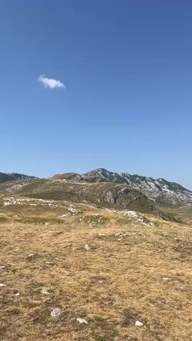 The video shows a vast mountainous landscape under a clear blue sky with a single white cloud. The foreground is dominated by dry, yellow grass and scattered white rocks. In the mid-ground, the terrain transitions to rolling hills covered in patches of green vegetation and more rocks, with some areas appearing bare and rocky. The background features a prominent mountain range with rugged peaks, some of which are capped with white, likely snow or lighter colored rock. The overall impression is of a remote, natural environment. The speaker in the audio is heard asking "How are you? Where are you?" and then "Where, damn it, are you?", indicating they are looking for someone.