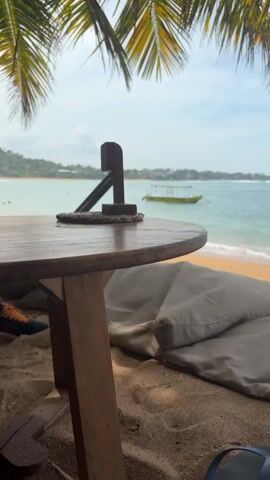 The video shows a tropical beach scene. In the foreground, there is a round wooden table with a coaster and a small wooden object on top. Next to it are several large, beige-colored cushions. The sand is golden brown. The view extends to the ocean, where several boats are anchored. One boat is labeled "SUN DIVING". The ocean is turquoise, and the sky is a mix of blue and white clouds. Palm tree fronds hang from the top of the frame, partially obscuring the sky. To the right, a sign displays text. In the distance, a glimpse of land with green vegetation can be seen. The camera slowly pans from left to right.
