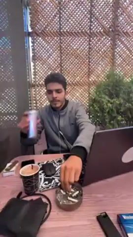 A young man sits at a table with a laptop, drinks from a can, and uses an electronic cigarette. The man is wearing a gray jacket. The table is cluttered with a coffee cup, an ashtray filled with cigarette butts, a mouse, and a pack of Winston cigarettes. He is wearing earphones. The background features a woven wall covering and greenery. He closes his eyes slightly and purses his lips after using the e-cigarette.
