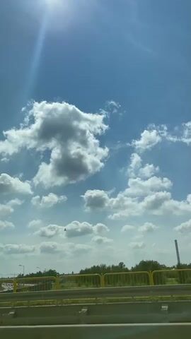 A vertical, full-shot video shows a bright, sunny day with a clear blue sky dotted with fluffy white clouds. The sun shines intensely in the upper left corner, casting a lens flare. Below the sky, a landscape features a highway with cars driving in the distance. A yellow-railed fence runs horizontally across the middle of the frame, separating the highway from the foreground. Trees and vegetation are visible behind the fence. A tall, slender structure, possibly a chimney or a pylon, stands in the background on the right. The overall impression is one of a pleasant, open outdoor environment.