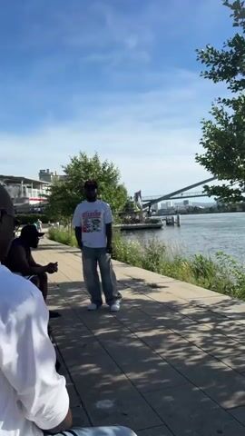 A man wearing a white t-shirt, light-colored pants, and a baseball cap stands on a paved walkway next to a body of water. A bridge is visible in the distance over the water. A young man wearing a white polo shirt, denim shorts, and sneakers walks past pushing a stroller. The scene appears to be outdoors on a sunny day with trees and buildings visible in the background.