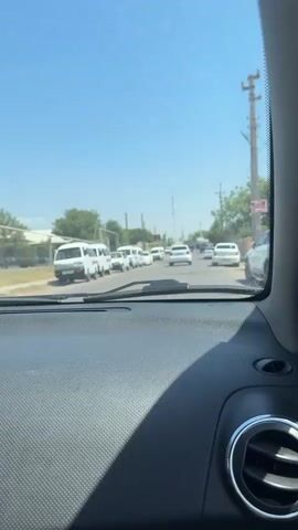 The video captures a view from inside a vehicle looking forward through the windshield. The vehicle appears to be moving along a street lined with parked cars and trees. The sky is clear and blue, indicating a sunny day. The road ahead has several vehicles, suggesting moderate traffic. The dashboard of the vehicle is visible in the foreground.