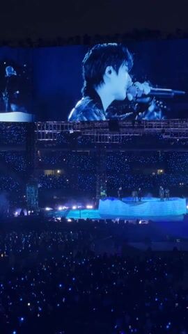 The video shows a live concert performance from a large audience's perspective. A male singer, wearing a dark jacket, performs on stage, singing into a microphone. A large screen above the stage shows a close-up of the singer's face as he sings. The stage, illuminated in blue light, has a multi-tiered platform where the singer and other performers are visible. The audience is packed in, with many holding up lights, creating a sea of blue dots in the dark stadium. The video focuses on the singer and the overall atmosphere of the concert.