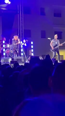 A band is performing on an outdoor stage at night in front of a crowd of people. The lead singer, a man with blonde hair, is wearing a black leather jacket and singing into a microphone. A guitarist, wearing a black tank top, stands to his right, playing a bass guitar. A drummer is visible behind the singer. The stage is lit with blue and white lights. The crowd is silhouetted against the stage lights, with many people holding up their phones to record the performance. The background shows a building with several windows.