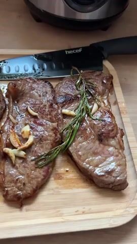 A close-up shot pans over four cooked steaks, garnished with rosemary sprigs and sliced garlic cloves, arranged on a light-colored wooden cutting board. A black-handled knife with a silver blade is positioned on the board to the right of the steaks. The steaks appear to be seasoned and seared, with visible juices pooling around them. The background is blurred but suggests a kitchen environment with a wooden countertop and a silver appliance, possibly a rice cooker, in the upper right corner. The lighting is bright, highlighting the texture and sheen of the meat.