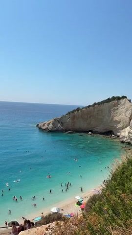 The video shows a vibrant beach scene with clear turquoise water and a bright blue sky. The beach is populated with many people swimming, sunbathing, and relaxing. Some individuals are floating on inflatable toys in the water, while others are wading near the shore. The coastline is characterized by a sandy beach, with rocky cliffs and lush green vegetation bordering the water. A few beach umbrellas and chairs are scattered across the sand. In the distance, a white yacht is visible on the water, and the coastline curves around a rocky promontory. The video is shot from a slightly elevated perspective, looking down onto the beach and the sea.