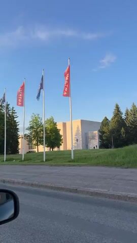 The video, filmed from inside a moving car in Imatra, Finland, captures a sunny outdoor scene.

The initial frames show several flags aligned along a road. Some flags are red with white text, partially visible as "SATRA," likely referring to "IMATRA." Other flags are blue with white text. Behind the flags, a modern, light-colored building is visible amidst green trees and grass.

As the car continues, a blue and white road sign comes into view on the right side of the road. The sign displays symbols for pedestrians and cyclists and appears to have Finnish text, possibly "KEUPAN TIE ALAKAAN" or similar, indicating a road name or direction.

The camera then shifts to show a row of lush green trees lining the road on the right, with a clear blue sky overhead. The rearview mirror of the car is prominent in the foreground, reflecting a person, likely the driver or passenger, holding a smartphone, suggesting they are filming the video.

The overall impression is a pleasant, clear day, with sunlight illuminating the landscape.