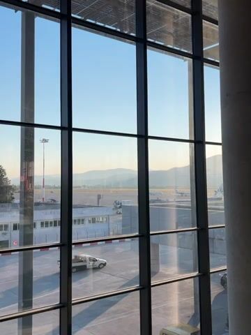 The video shows an airport tarmac viewed through a large window. In the foreground, a white utility truck is parked on the tarmac. In the midground, there are several airport buildings and vehicles, including what appears to be an airplane in the distance. The background features a hazy landscape of mountains under a clear blue sky. The sun is shining, casting long shadows on the tarmac. The overall impression is a quiet airport scene in the morning or late afternoon.