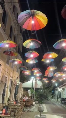 The video shows a vibrant street scene at night in Karditsa, Greece. Numerous colorful, rainbow-patterned umbrellas are suspended across the street overhead, each illuminated by a light bulb, creating a decorative canopy. The street is flanked by buildings on both sides. On the left, an outdoor cafe area is visible with tables and wicker chairs, where several people are seated, including a man in a light-colored shirt. Further down the street, another outdoor seating area can be seen on the right. A scooter briefly passes in the background. The overall atmosphere is lively and inviting, enhanced by the unique umbrella display.