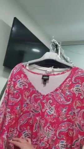 A woman with blonde hair stands in front of a rack of clothing. She is wearing a yellow shirt. The rack contains various garments in different colors, including a pink paisley top, a black garment, a bright pink garment, a purple garment, and a blue garment. A television is visible in the background.