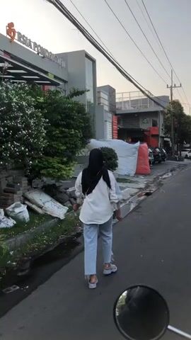 The media file depicts a person walking down a street in Pagerwojo, East Java. The individual, seen from behind, is wearing a white shirt, light blue jeans, and a black hijab. They are also wearing white sandals.

The scene appears to be an urban or semi-urban environment with a paved road. To the left of the road, there's a sidewalk with some debris, including sacks and what looks like discarded building materials, as well as a lush green bush with white flowers. Further back, a modern building with large windows is visible, and signage indicates it is "RSIA PONDOK GUCI." Adjacent to this building, there's another structure with red and white elements. Across the street, more buildings and parked cars are present, along with overhead utility wires.

The time of day appears to be late afternoon or early evening, suggested by the warm, soft lighting and the long shadows cast by the objects. The weather seems calm, with no indication of rain or strong winds.

The overall mood conveyed is casual and ordinary, a person going about their day. The presence of a motorcycle mirror in the bottom right corner suggests the recording was potentially made from a motorcycle. The visible text "RSIA PONDOK GUCI" provides a specific location cue, identifying a hospital.