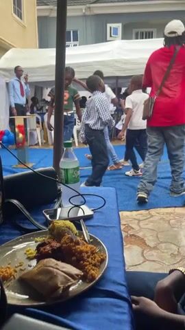 The video shows an outdoor event with a gathering of people, predominantly children, dancing on a blue carpeted area under a tent. In the foreground, a plate of food with rice, meat, and some sort of wrap is visible on a blue tablecloth. A bottle of Sprite and a phone are also on the table. Several children are actively dancing in the center of the frame, dressed in casual and semi-formal attire. Adults are present in the background, some observing the children and others possibly involved in organizing the event. A building with light-colored walls and windows is visible behind the tent. The overall atmosphere appears to be festive and lively.
