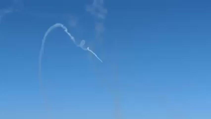 The video shows an airplane performing aerobatics in a bright blue sky, leaving a white smoke trail. The camera pans down to reveal the ocean, a crowded beach, and buildings along the coastline. Two yellow boats are visible on the water. The scene is outdoors, likely a public event or air show over the ocean.