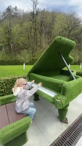 A young girl with blonde hair, wearing sunglasses, a light-colored, possibly tie-dye jacket, and light blue jeans, is seated on a grass-covered bench outdoors. She is playfully interacting with a large, artfully crafted grand piano sculpture that is entirely covered in vibrant green artificial turf. Her hands are positioned on the white and black keys, simulating playing the instrument. The piano's lid is propped open, revealing more of the green turf interior.

The scene is set on a bright, sunny day with clear skies. The ground beneath the piano is paved with light-colored tiles, and a metal drain grate is visible in the foreground to the right. The bench the girl sits on has a visible brown wooden base. In the background, an extensive, well-manicured green lawn stretches out, bordered by a dense forest of lush deciduous trees covering rolling hills or mountains. The natural, verdant backdrop complements the unique grass art installation. This whimsical setting is consistent with the beautiful natural landscapes found in Большой Сочи, Russia. No visible text is present in the image.