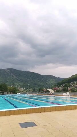 A wide outdoor swimming pool with multiple lanes is visible. The water is blue with yellow and pink lane markers. The pool is bordered by a beige tiled area. In the background, there are green, tree-covered hills and some buildings. The sky is cloudy. There are a few people in the pool, and some lounge chairs on the side. The camera is static.