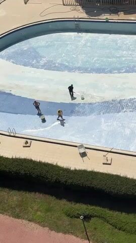 The video, filmed from an elevated perspective, captures a large, empty swimming pool in Erdemli, Turkey, undergoing maintenance. The pool floor is covered with light and dark blue patterned tiles.

Three men are actively working inside the pool. Each worker uses a long-handled brush or squeegee, appearing to spread or remove a substance across the tiled surface, which creates streaks and highlights the tile patterns. Buckets are placed on the pool floor near each individual. As the camera pans, a fourth worker is visible near the pool's edge.

The pool deck, made of light-colored paving stones, surrounds the pool. Pool ladders are present on the sides of the deep end. A wheelbarrow and other tools are visible on the deck. A neat, low hedge borders the pool area, separating it from a grassy section. In the background, parts of a structure, possibly a building or equipment related to the pool, are discernible.

The scene is brightly lit by natural daylight, casting clear shadows. The activity suggests a thorough cleaning, restoration, or preparation of the pool before it is refilled. No visible text is present in the video.