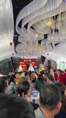 This video captures an indoor wedding reception or ceremony taking place in Bình An, Vietnam. The venue is festively decorated with white fabric swags and yellow hanging lanterns on the ceiling, along with blue and white fairy lights.

On a raised stage, the bride, dressed in a white wedding gown and holding a bouquet, stands alongside the groom, who is wearing a dark suit. Behind them, a large LED screen displays red heart graphics and Vietnamese text, which partially reads "Lễ Tân Hôn" (Wedding Ceremony), and below it, the names "Quang" and "Ngọc Ý," likely identifying the couple. To the right of the couple, a person in a yellow traditional Vietnamese outfit (Áo Dài) is standing, possibly a host or singer, with another individual seated at a keyboard nearby. Another person in a similar yellow outfit is visible to the left of the couple.

Numerous guests are seated at tables in the foreground and midground, observing the proceedings on stage. Some guests are focused on the stage, while others are engaging in conversation or looking towards the camera. One guest is seen holding up a phone to record the event. The ambiance is celebratory, indicative of a wedding.
