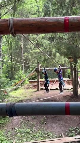 In a forest setting, two individuals are navigating an outdoor adventure course. They are suspended by safety harnesses and are walking on a thin rope bridge, with a large wooden log positioned above them. The individuals are wearing helmets and appear to be children. One child is wearing a dark blue outfit with black leggings, and the other is in a purple and blue outfit with black leggings. They are both holding onto cables or ropes for support.

The scene is set amongst tall trees with lush green foliage, suggesting a daytime scene in warm weather, possibly summer or late spring. The ground beneath them is covered with dirt and scattered leaves. There are other ropes and wooden structures visible in the background, indicating a well-established adventure park or obstacle course.

The overall mood seems to be one of focused activity and possibly a bit of trepidation as the children make their way across the suspended walkway. The presence of safety equipment suggests an organized activity designed for recreation and challenge. The visual information does not contain any discernible text. The location is identified as Sergino, Russia.