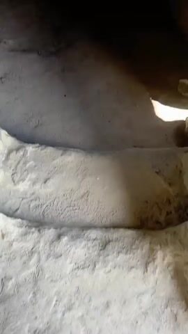 The video shows the interior of a small cave-like space. The walls are made of rough, light-colored stone, with evidence of erosion and chipping. The floor is dusty and uneven. Light enters from an opening, creating a contrast between light and shadow within the space. The focal point of the space is a small alcove decorated with several framed religious icons and paintings. Two larger icons are prominently displayed on the floor. There is no visible human interaction in the video.