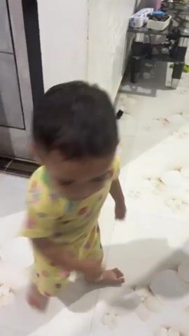 A baby in a yellow outfit with colorful patterns is walking around and playing. The baby appears to be exploring their surroundings, moving from one area to another. The floor has a tiled pattern with floral designs. There are also some white objects scattered on the floor. The background includes a brown curtain and a light-colored wall. The video captures the baby's playful movement as they interact with the environment.