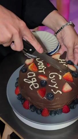 A close-up shot focuses on a round, chocolate-covered cake adorned with strawberries and blueberries. A person's hands are visible, one holding a black-handled knife as it cuts into the cake. White frosting spells out text on the cake, which appears to read "Don't Age Hot Guys". The cake sits on a gray plate, which rests on a metallic surface. In the background, a person's wrist wearing a beaded bracelet and a watch is visible. A white, ridged object is also present in the background. The lighting suggests an indoor setting, possibly at night.