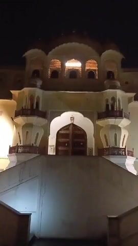 The video shows a still shot of a large, ornate building at night. The building is mostly light yellow in color with white accents and is illuminated by lights from within. The central part of the building features a large arched doorway with wooden doors that are closed. On either side of the doorway are balconies with detailed railings. Above the doorway, the building has an upper level with a row of latticed windows and decorative domes on top. A broad set of white steps leads up to the building's entrance. The sky is dark, indicating it is nighttime.